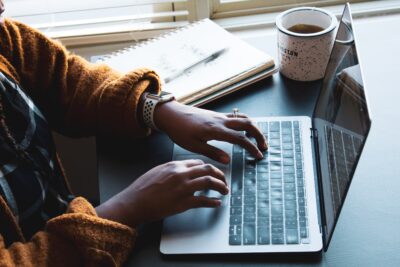 hands typing on a laptop computer keyboard. A notepad and coffee cup sit nearby